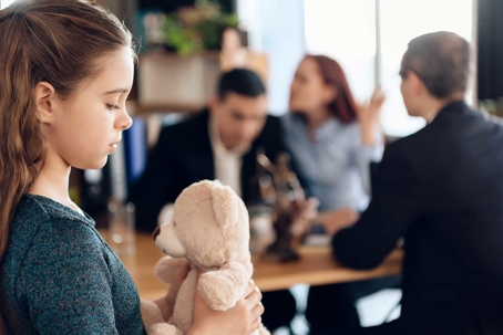 a child holding a teddy bear in front of her fighting parents.